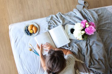 Gri yatakta servis edilen kruvasan ve kahveyle kahvaltı. Güzel genç kız yatakta uzanıyor ve kitap okuyor..