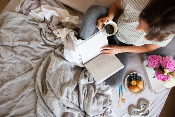 Gri yatakta servis edilen kruvasan ve kahveyle kahvaltı. Güzel genç kız yatakta uzanıyor ve kitap okuyor..