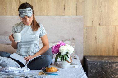 Gri yatakta servis edilen kruvasan ve kahveyle kahvaltı. Güzel genç kız yatakta uzanıyor ve kitap okuyor..