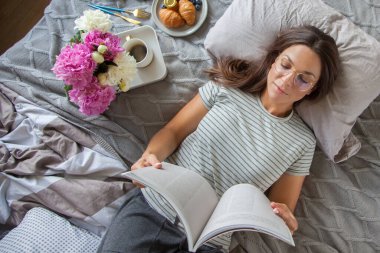Kitap ya da gazete okuyan ve sabahları yatakta kahve içen bir kadın.