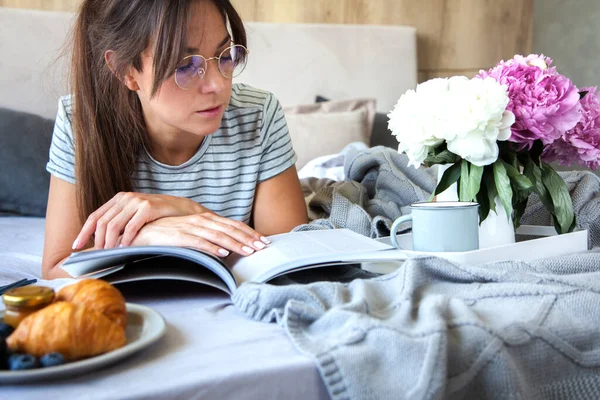 早餐与羊角面包和咖啡供应在灰色的床上 美丽的小女孩躺在床上看书 — 图库照片
