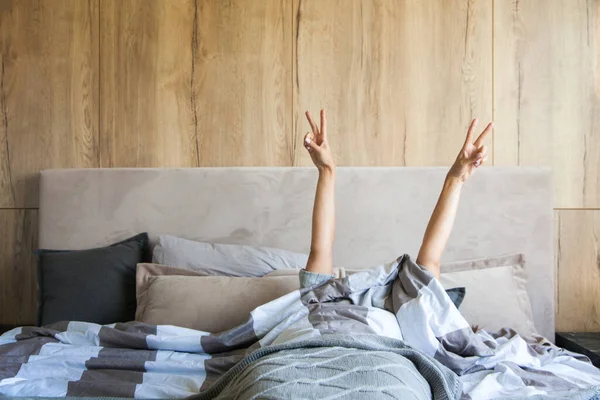Young woman waking up in the morning in the bed, hidding under the blanket, stretching out one\'s arms with a cup of coffee and showing peace sign