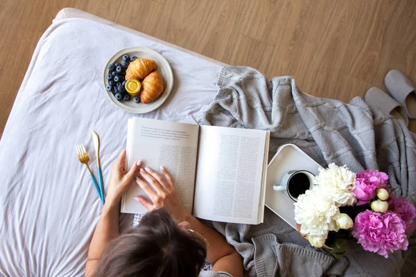 Gri yatakta servis edilen kruvasan ve kahveyle kahvaltı. Güzel genç kız yatakta uzanıyor ve kitap okuyor..