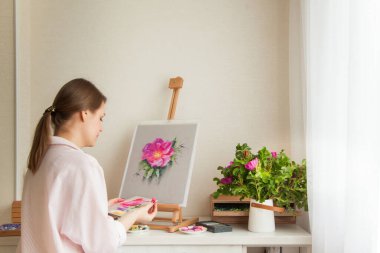 Stüdyoda çalışan, gülümseyen genç, pozitif bir sanatçı sehpası ve buketi olan pembe gül çiçeğiyle iş için yatağın renklerini seçiyor. Yaratıcılığı meslek olarak algıla