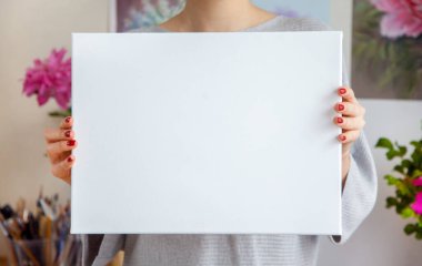 Gülümseyen genç kadın sanatçı, ellerinde boş beyaz bir tuval, duvarında resimler ve aletlerle rahat bir iş yerinin arka planına dayanıyor. Reklam alanı