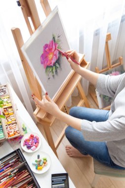 Genç kadın ressamın arka görüntüsü, açık hava ahşap bir sehpada otururken atölyesinde kuru pastel boya kalemleri olan bir kutudan pembe şakayık resmi çiziyor. Yaratıcılık ve hobi konsepti. Pembe gölgeler.