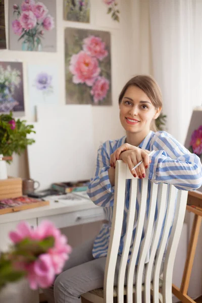 Caucasian beautiful smiling girl artist with glasses paints in home studio on background of pink flowers and paintings on wall with craftsmanship for creativity. Creativity and inspiration concept