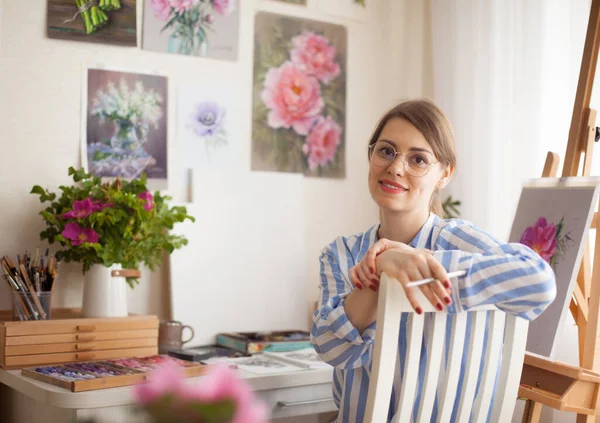 Caucasian beautiful smiling girl artist with glasses paints in home studio on background of pink flowers and paintings on wall with craftsmanship for creativity. Creativity and inspiration concept