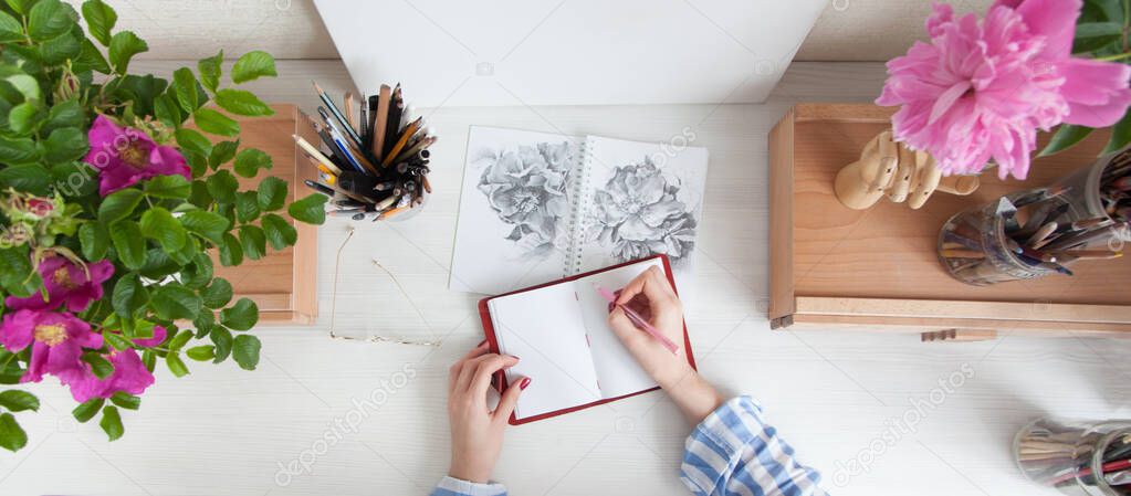 Top view girl artist draws a sketch of rose hip flowers sitting at her workplace with bouquets of flowers and materials for creativity. Hobby and creation concept