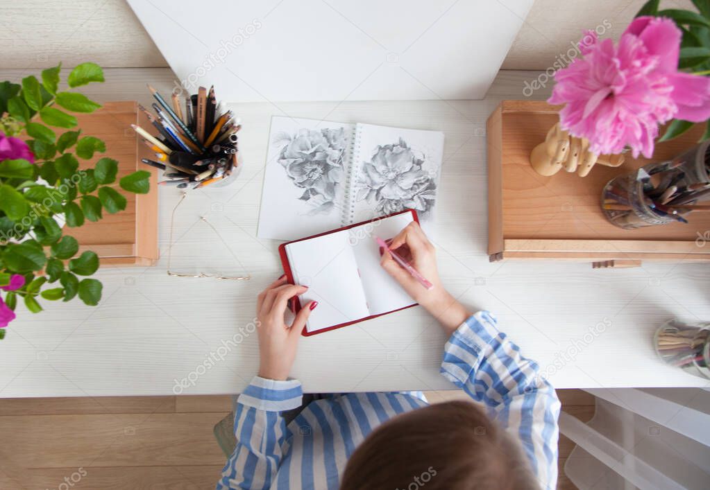 Top view girl artist draws a sketch of rose hip flowers sitting at her workplace with bouquets of flowers and materials for creativity. Hobby and creation concept