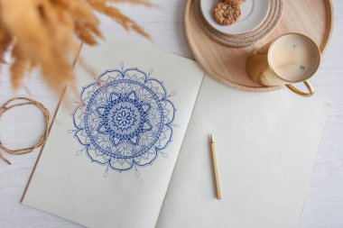 Bir kadın beyaz bir çarşafa karalama mandala çiziyor. Rahat ol. Beyaz ahşap masa. Yıllanmış ahşap. Bir kupa kahve ve çikolatalı kurabiye. Tespih ve ahşap boncuklar.