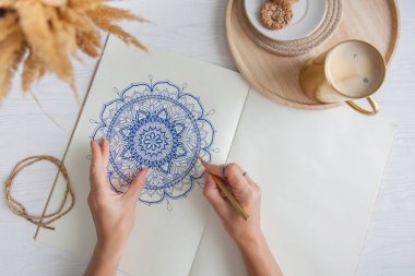 Bir kadın beyaz bir çarşafa karalama mandala çiziyor. Rahat ol. Beyaz ahşap masa. Yıllanmış ahşap. Bir kupa kahve ve çikolatalı kurabiye. Tespih ve ahşap boncuklar.