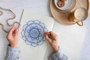 Bir kadın beyaz bir çarşafa karalama mandala çiziyor. Rahat ol. Beyaz ahşap masa. Yıllanmış ahşap. Bir kupa kahve ve çikolatalı kurabiye. Tespih ve ahşap boncuklar.