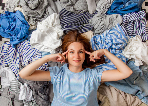 Beautiful caucasian woman smiling and lying down with clutter clothes on the floor. Decluttering pile of messy wardrobe. Cluttered housewife. Fabric. Shopping. Housework idea concept.