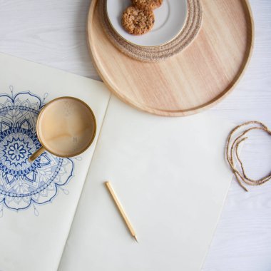 Bir kadın beyaz bir çarşafa karalama mandala çiziyor. Rahat ol. Beyaz ahşap masa. Yıllanmış ahşap. Bir kupa kahve ve çikolatalı kurabiye. Tespih ve ahşap boncuklar.