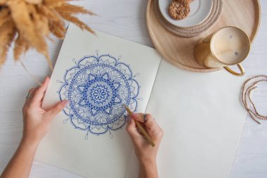 Bir kadın beyaz bir çarşafa karalama mandala çiziyor. Rahat ol. Beyaz ahşap masa. Yıllanmış ahşap. Bir kupa kahve ve çikolatalı kurabiye. Tespih ve ahşap boncuklar.