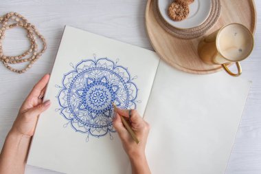 Bir kadın beyaz bir çarşafa karalama mandala çiziyor. Rahat ol. Beyaz ahşap masa. Yıllanmış ahşap. Bir kupa kahve ve çikolatalı kurabiye. Tespih ve ahşap boncuklar.