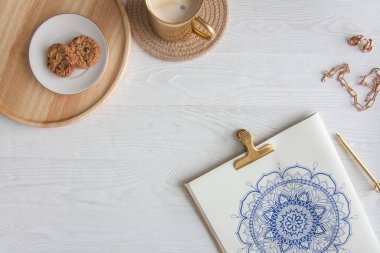 Bir kadın beyaz bir çarşafa karalama mandala çiziyor. Rahat ol. Beyaz ahşap masa. Yıllanmış ahşap. Bir kupa kahve ve çikolatalı kurabiye. Tespih ve ahşap boncuklar.