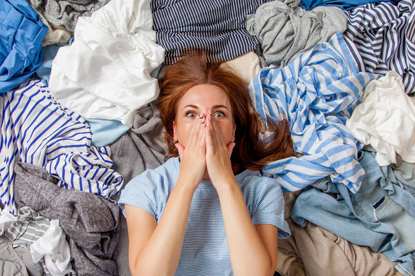 Beautiful caucasian woman smiling and lying down with clutter clothes on the floor. Decluttering pile of messy wardrobe. Cluttered housewife. Fabric. Shopping. Housework idea concept.