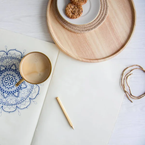 Bir kadın beyaz bir çarşafa karalama mandala çiziyor. Rahat ol. Beyaz ahşap masa. Yıllanmış ahşap. Bir kupa kahve ve çikolatalı kurabiye. Tespih ve ahşap boncuklar.