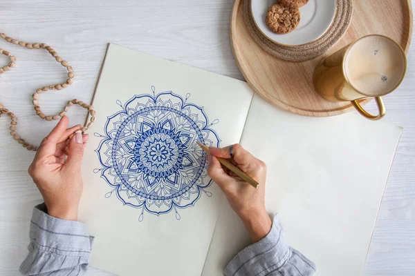 Bir kadın beyaz bir çarşafa karalama mandala çiziyor. Rahat ol. Beyaz ahşap masa. Yıllanmış ahşap. Bir kupa kahve ve çikolatalı kurabiye. Tespih ve ahşap boncuklar.