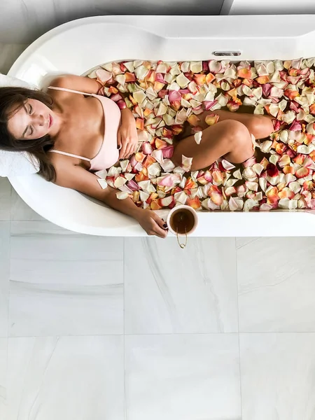 年轻漂亮的姑娘躺在有粉色花瓣的浴缸里 从上面看喝粉红色杯子里的茶 在一个大的圆形的白色浴缸里 — 图库照片