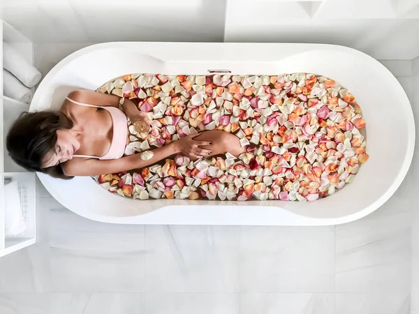 Genç ve güzel bir kız pembe yapraklarla bir banyoda yatıyor. Yukarıdan bak. Pembe bardaktan çay içiyor. Büyük yuvarlak beyaz bir küvette.