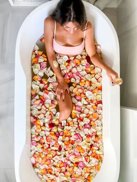 若い美しい少女はピンクの花びらのバスルームにあります 上からの眺め ピンクのマグカップからお茶を飲む 大きな丸い白い浴槽で — ストック写真