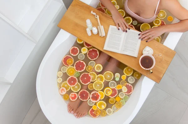 Top view of faceless female in bathtub with slices of oranges, lemon and grapefruits is reading the book. Woman having detox bath treatment. Pedicure tools on wooden tray. Relaxing concept. Vacation.