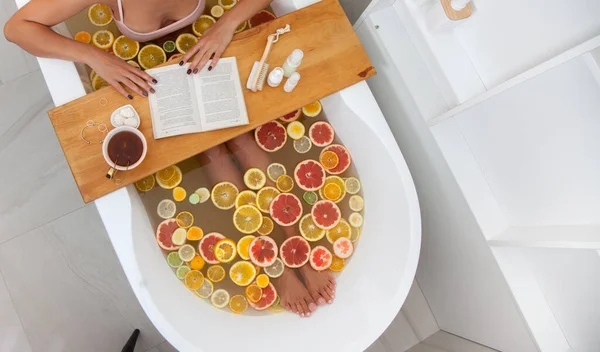在浴缸里 带着橙子 柠檬和柚子片的无脸女性的头像正在阅读这本书 正在接受排毒洗浴治疗的妇女 木制托盘上的教学工具 放松的概念 — 图库照片