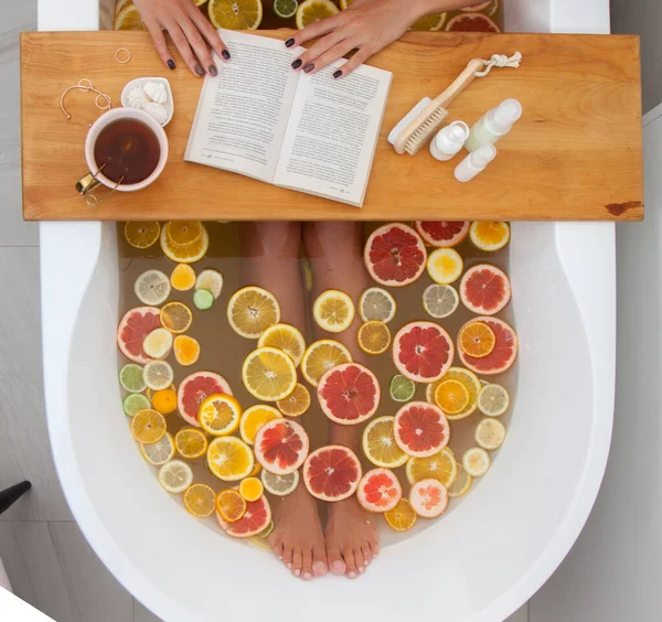 Top view of faceless female in bathtub with slices of oranges, lemon and grapefruits is reading the book. Woman having detox bath treatment. Pedicure tools on wooden tray. Relaxing concept. Vacation.