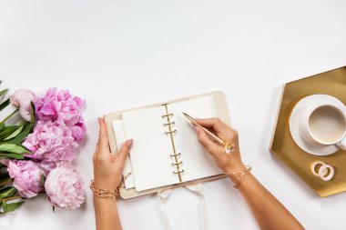Mücevher içindeki iyi giyimli kadınlar günlüğüne aletler, ofis malzemeleri seti, yanında bir fincan kahve ve beyaz arka planda şakayık buketi ile birlikte yazıyorlar. Girişimci çalışma alanı.
