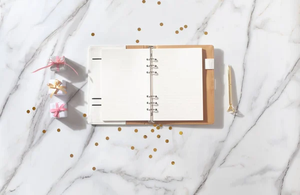 Notebook with empty pages for your text, pen, coffee cup, milk, small gift boxes with pink ribbon and golden sparkles on white marble table. New Year and Christmas concept, top view. Festive holidays