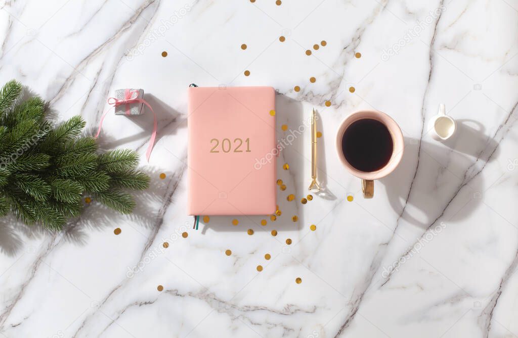 Flat lay composition with coral colored 2021 diary book for writing down New Year's plans, coffee, cookie and mini artificial Christmas trees onside with gold sparkles on white background. Holiday