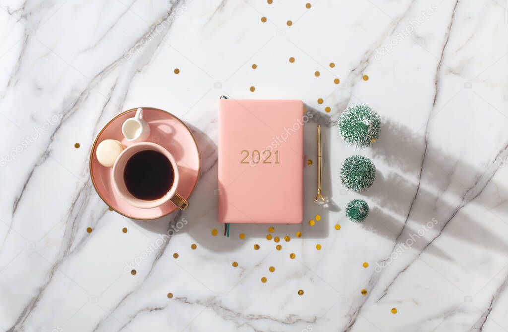 Flat lay composition with coral colored 2021 diary book for writing down New Year's plans, coffee, cookie and mini artificial Christmas trees onside with gold sparkles on white background. Holiday