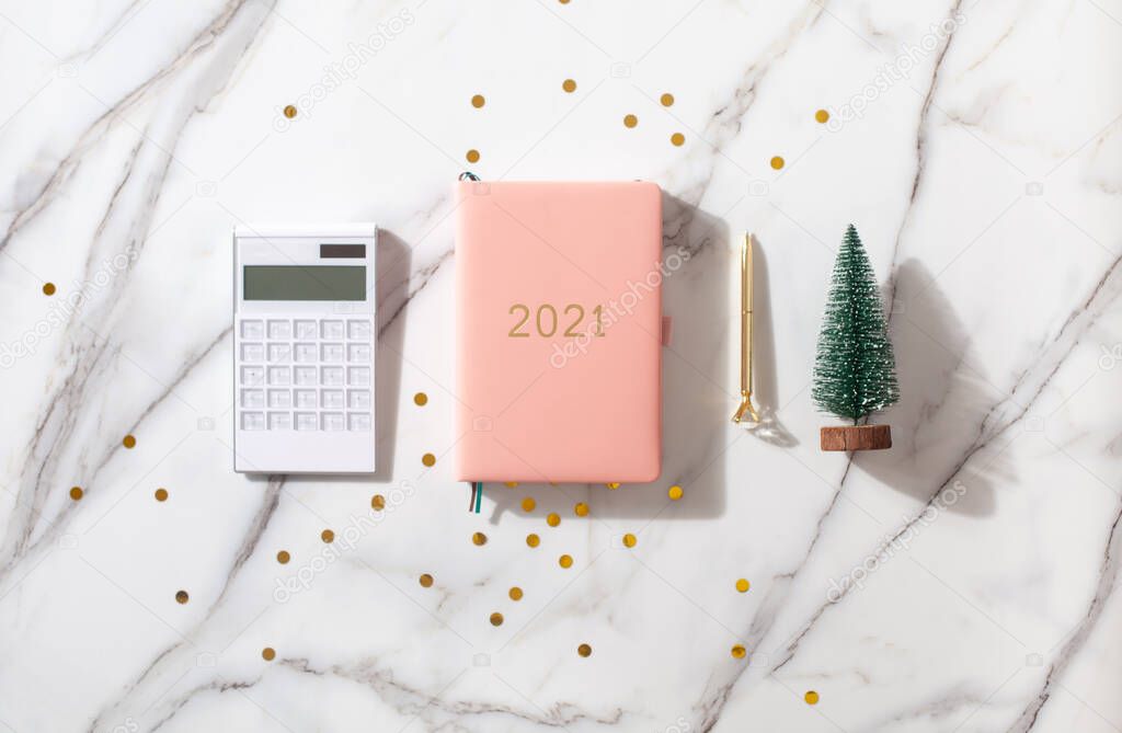 Flat lay composition with coral colored 2021 diary book for writing down New Year's plans, coffee, cookie and mini artificial Christmas trees onside with gold sparkles on white background. Holiday