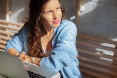 Mavi örgülü, uzun saçlı mutlu genç kadın pencerenin yanında oturuyor ve uzaktan bilgisayarla çalışıyor. Pencere manzaralı bir ev. Zarif iş kadını gülümsüyor..