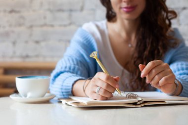 İyi giyimli kadın elinde altın kalemle not defterine altın kalemle notlar yazarken pencerenin yanında mavi latte içiyor. Evde çalışan serbest çalışan bir gazeteci. Gelecek planlama konsepti. Boşluğu kopyala