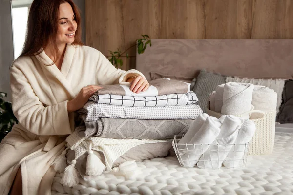 Kışın kalın ve kalın bornozlu güzel bir kadın oturmuş çarşafları düzgünce katlıyor ve beyaz banyo havluları kullanıyor. Temiz çamaşırları düzenlemek ve sıralamak. Organik ve doğal pamuk tekstili. Üretim.