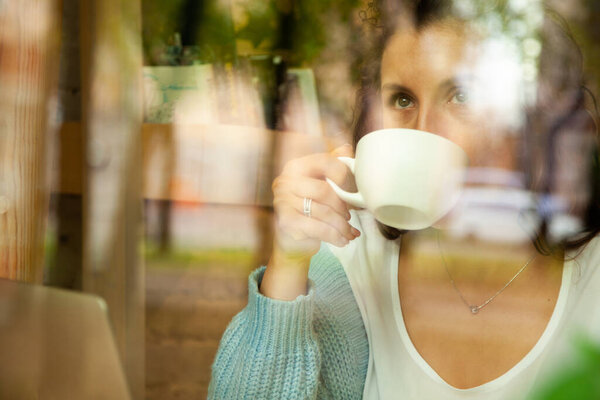 View through window of young woman wearing blue sweater talking on phone while working on computer and enjoying cup of hot drink in coffee shop. Street reflection. Freelance concept.