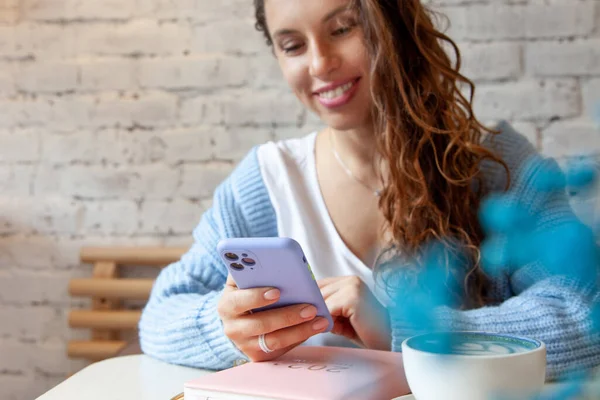 身穿蓝色保暖毛衣的年轻女子边喝咖啡边看手机短信 女商人正在用她的智能手机查看社交媒体 博客生活方式 规划和组织工作 — 图库照片