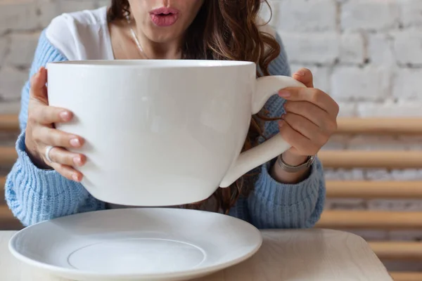 Young woman in blue sweater frowning with head in hand and holding giant cup of tea at home. Woman is feeling headache, holding big cup hot drink. Concept of disease, sickness and illness. Copy space