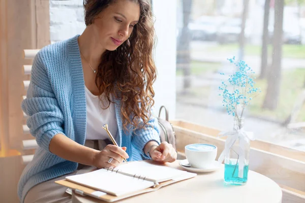 Mavi, sıcak kazaklı genç bir kadın büyük bir kahve dükkanının penceresinde oturuyor ve mavi latteyle bir Noel alışveriş listesi yazıyor. Noel tatili planlıyorum. Düzenleme ve planlama konsepti.