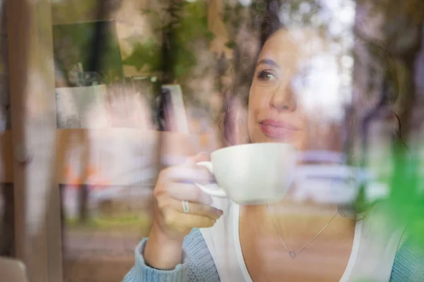 透过窗户 可以看到年轻女子穿着蓝色毛衣 一边打电话一边在电脑上工作 一边在咖啡店里喝着热饮的情景 街头反思 自由的概念 — 图库照片