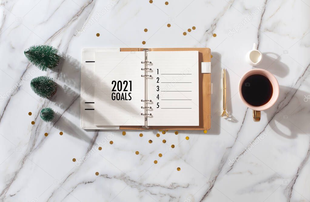 Flat lay composition with coral colored 2021 diary book for writing down New Year's plans, coffee, cookie and mini artificial Christmas trees onside with gold sparkles on white background. Holiday