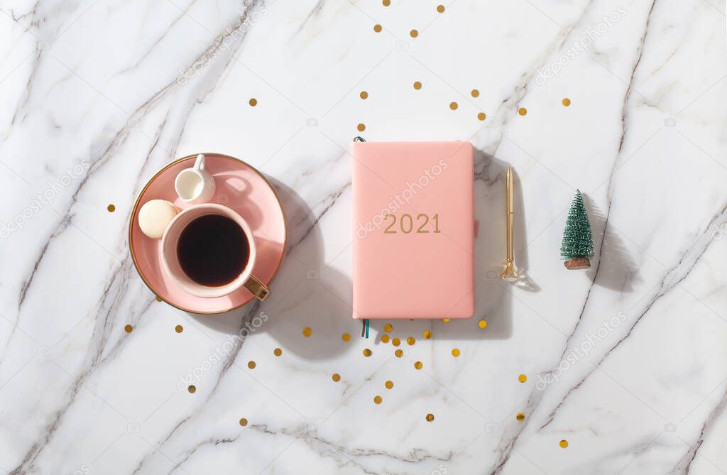 Flat lay composition with coral colored 2021 diary book for writing down New Year's plans, coffee, cookie and mini artificial Christmas trees onside with gold sparkles on white background. Holiday