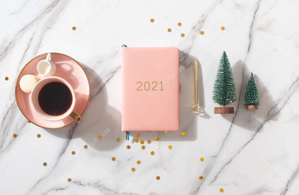 Flat lay composition with coral colored 2021 diary book for writing down New Year's plans, coffee, cookie and mini artificial Christmas trees onside with gold sparkles on white background. Holiday