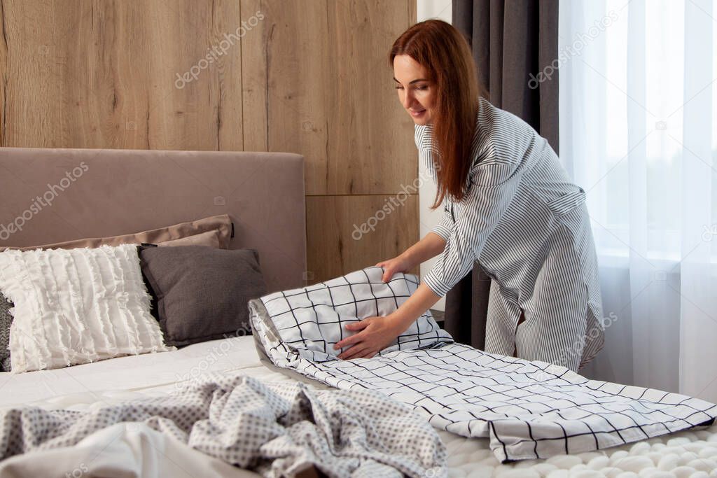 Happy woman with long red hair is folding bedding set on the bed of room with copy space. Organic textile manufacture. Linen are decorated with stripes in the form of a cage. Different pillows
