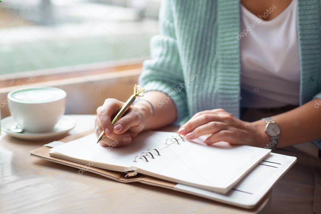 Well groomed woman hand holding gold pen and writing notes with gold pen in notebook while drinking blue latte beside window. Freelance journalist working at home. Planning future concept. Copy space