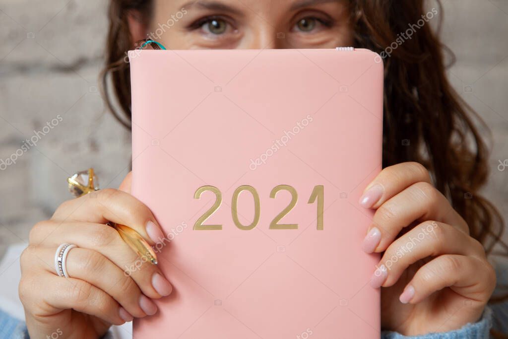 Woman with wavy hair is closing her face with a pink coloured planner 2021 near beige brick wall. New year planning concept with future ideas and goals. Copy space